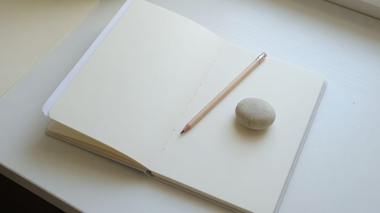 자연광 아래 흰 책상 위에 펼쳐진 빈 노트와 신중하게 놓인 연필 하나, 작은 조약돌 하나가 함께 있는 에디토리얼 정물 이미지 — 시험을 앞두고 마음을 고르는 차분한 수험생 책상 구도